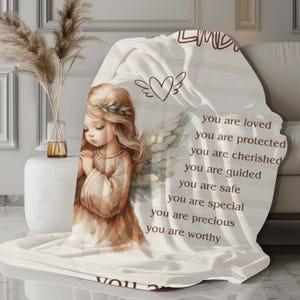 May include: A white throw blanket with a watercolor illustration of an angel with wings and the text "You are loved, you are protected, you are cherished, you are guided, you are safe, you are special, you are precious, you are worthy."