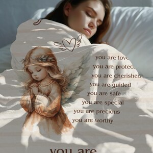 May include: A white blanket with a brown angel illustration and the text "you are love, you are protected, you are cherished, you are guided, you are safe, you are special, you are precious, you are worthy, You are" printed on it.