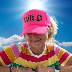 May include: A woman wearing a pink trucker hat with the word "WILD" in black and leopard print letters. She is wearing a striped shirt with a rainbow pattern.