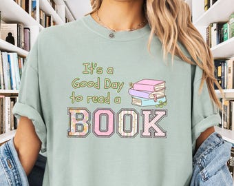 May include: A light green t-shirt with the text "It's a Good Day to read a BOOK" in pink and green letters. The word "BOOK" is decorated with floral patterns. A stack of books with floral patterns is illustrated above the word "BOOK".