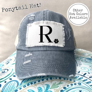 Może przedstawiać: Niebieski denimowy kapelusz z warkoczem z białą naszywką z napisem "R." Kapelusz ma siateczkowe plecy i przetarty wygląd. Dostępne są inne kolory kapeluszy.