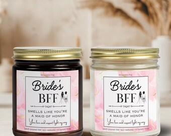 Könnte beinhalten: Zwei gläserne Kerzenbehälter mit goldenen Deckeln und rosa-goldenen Etiketten. Die Etiketten lauten "Bride's BFF" und "Smells like you're a maid of honor. Your love and support light my way." Die Kerzen sind handgefertigt in den USA und bestehen aus natürlichen Zutaten.