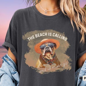 Może przedstawiać: Szary t-shirt z nadrukiem buldoga w okularach przeciwsłonecznych i kapeluszu. Tekst na koszulce brzmi "The beach is calling and I must go".