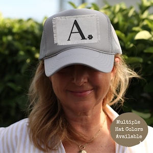 Może przedstawiać: Szara czapka trucker z białym naszywką z napisem "A." i małym czarnym kwiatkiem obok litery. Dostępne są różne kolory czapek.