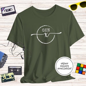May include: A green t-shirt with a white circle graphic that says "GEN X" in a handwritten font. The text "other colours available!" is written in a white circle at the bottom of the shirt.