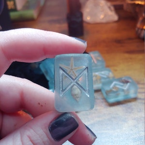 May include: A clear, rectangular resin rune with a silver symbol, a starfish, and seashells. The rune is held in a hand, with other similar runes in the background. A lit candle and other objects are visible.
