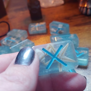 May include: Close-up of a blue resin dice with a white starfish and a blue X inside. The dice is being held by a hand.