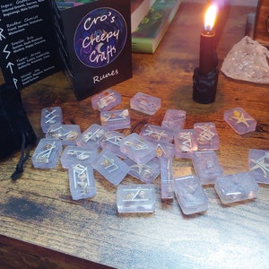 May include: A set of 25 clear resin runes with silver runes and embedded seashells. The runes are scattered on a wooden surface with a black velvet bag, a black candle, and a small wooden box with a pentagram design.