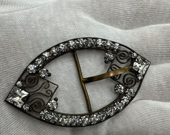 May include: A silver-toned metal buckle with a decorative design and clear rhinestones. The buckle is oval-shaped and has a T-shaped closure.