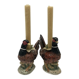 May include: A pair of ceramic pheasant candle holders, each featuring a hand-painted bird with a black head, red face, and brown body. Each pheasant stands on a textured base and holds a tall, cylindrical, light yellow beeswax taper candle.