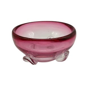 May include: A small, round, glass bowl with a deep magenta rim and a clear base. The bowl has three decorative feet and a smooth, glossy finish. The interior of the bowl is a lighter shade of pink.