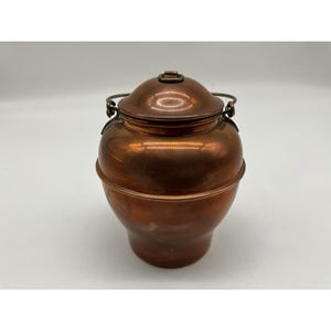 May include: A polished copper container with a rounded body and a matching lid. The vessel features a thin, curved metal handle attached to the sides with decorative loops. The surface has a warm, metallic sheen and a smooth finish.