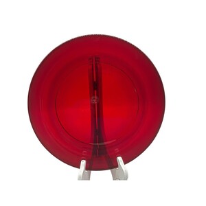 May include: A circular, translucent ruby red glass plate featuring a smooth, polished surface and a distinct central circular indentation. The plate is supported by a white stand, and a small circular logo is visible in the center of the glass.