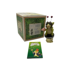May include: A Pocket Dragons Commander Cookie figurine featuring a light green dragon wearing brown aviator goggles and holding a pink object, standing on a black and white cookie base, with its original green box and a small illustrated card.