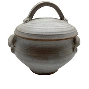 May include: A round, light gray ceramic lidded pot with a speckled texture. The pot features two side handles and a single arched handle on the lid. The base and the rim where the lid meets the pot show a reddish-brown clay color.