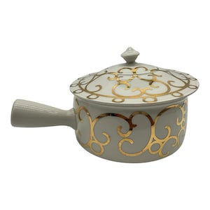May include: A light gray ceramic saucepan with a matching lid, featuring a prominent gold filigree scroll pattern on both the body and the lid. The pot includes a single side handle and a small, pointed knob on the center of the lid.