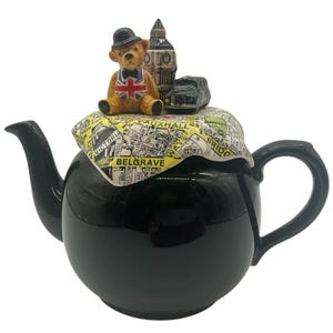 May include: A black ceramic teapot with a lid featuring a London-themed design. The lid displays a map of London, a brown teddy bear wearing a Union Jack vest and a black bowler hat, a miniature Big Ben clock tower, and a black London taxi cab.