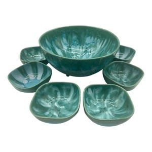 May include: A teal-colored ceramic serving bowl set. The set includes a large round bowl and six smaller square-shaped bowls. The bowls have a glossy finish with a marbled effect, showcasing shades of teal and white.