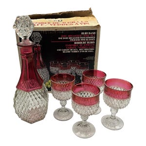 May include: A vintage Ruby Band wine set, including a decanter with stopper and four wine glasses. The decanter and glasses feature a clear, diamond-cut design with ruby red rims. The original box is visible.