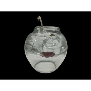 May include: A clear glass oil lamp with a round, bulbous shape. The lamp features a wick and a small, internal glass chamber. A small, oval sticker is visible on the front. The lamp is designed to hold oil for burning.