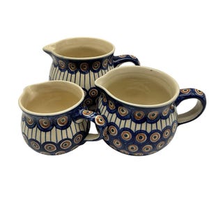 May include: Three ceramic pitchers with a blue and white pattern. The pitchers have a cream-colored interior and a handle. The design includes brown and blue circular and vertical stripe patterns. The pitchers are of varying sizes.