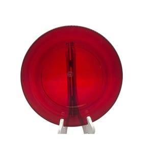 May include: A round, ruby red glass plate with a clear, vertical support stand. The plate has a smooth surface and a slightly raised rim. The stand is white and holds the plate upright. The plate is a decorative item.