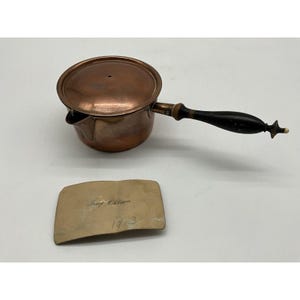 May include: A round copper saucepan with a lid and a long, dark wooden handle. Beside the pan lies a rectangular brass plaque engraved with the text Josef Ohlson and the year 1913. The copper surface shows a warm, metallic finish.