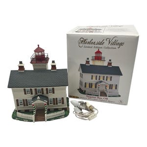 May include: A Harborside Village Yaquina Bay, OR porcelain lighted house with a white exterior, dark gray roof, and red lighthouse tower. The set includes a white cardboard box with product text and a white electrical cord with a light bulb.