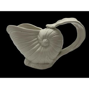 May include: A white ceramic gravy boat shaped like a nautilus shell. The vessel features a spiraled, ribbed exterior texture, a wide pouring spout, and a curved handle that extends from the top of the shell. The base is textured and solid.