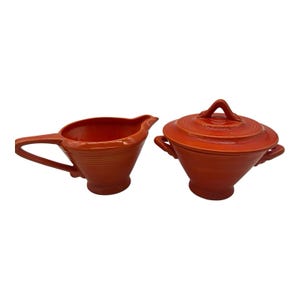 May include: A bright orange ceramic creamer pitcher and a matching lidded sugar bowl. Both pieces feature a distinct ribbed design around the body, a pedestal base, and smooth, glossy finish. The sugar bowl has two side handles and a lid.