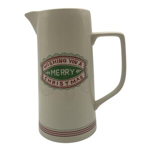 May include: Cream-colored ceramic pitcher with a rectangular handle and a spout. The pitcher features a red and green oval design with the text "Wishing You a Merry Christmas." A red dotted line encircles the base.