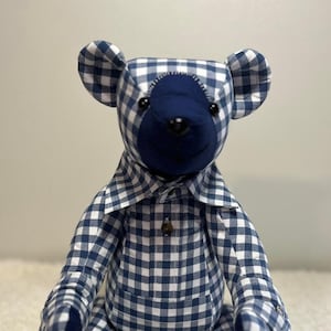 Könnte beinhalten: Ein blauer und weißer Gingham-Teddybär mit blauem Gesicht und Knopfaugen. Der Bär trägt ein Gingham-Hemd mit Knopfleiste. Die Pfoten des Bären haben die Daten "02.97" und "02.04.23" aufgestickt.