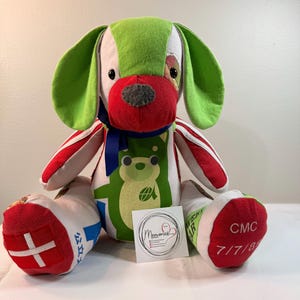 Pode incluir: Um cão de peluche feito de vários retalhos de tecido colorido, com orelhas verde-brilhantes, um nariz vermelho e uma fita azul à volta do pescoço. O corpo apresenta um gráfico de urso verde, um pé vermelho com uma cruz branca e o texto CMC 7/7/89.