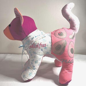 以下が含まれることがあります： ピンクと白のパッチワークで作られた猫のぬいぐるみで、胸に「Misifus」という名前が刺繍されています。おもちゃの足には花柄、しっぽにはピンクと白のドット柄が施されています。