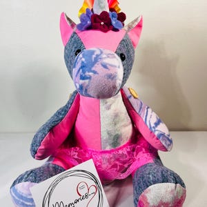 May include: A handmade unicorn plush toy in pink and grey. The unicorn has a pink face and a white horn. It is wearing a pink lace tutu and a floral crown. A tag with the word "Memories" is in front of the toy.