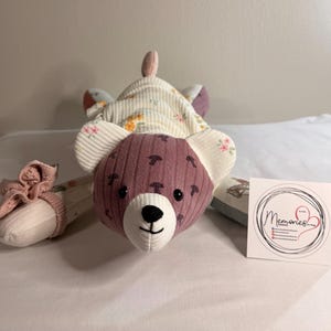 Pode incluir: Um urso de pelúcia feito à mão com uma cabeça roxa e orelhas e corpo com estampa floral. O urso tem um nariz preto e uma boca sorridente. Um laço rosa combinando está preso a um dos pés do urso. Um pequeno cartão com a palavra "Memories" está ao lado do urso.