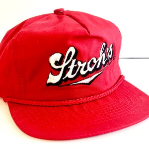 May include: A red baseball cap with the embroidered text "Stroh's" in white script with black accents. The cap features a braided rope detail along the brim and a small red button on top. The hat is a classic style.