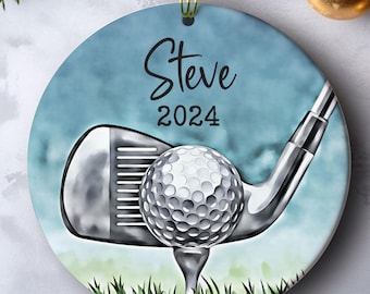 May include: Round ceramic ornament with a golf theme. The ornament features a golf club, golf ball, and the name "Steve" with the year "2024" on a blue background. The ornament is hanging with a gold string.