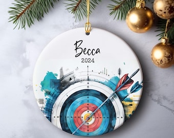 May include: A white ceramic ornament with the name "Becca" and the year "2024" in black script. The ornament features a colorful archery target design with a red and blue arrow. Gold hanging loop.