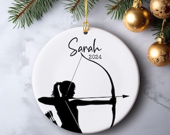 May include: White ceramic ornament with a black silhouette of an archer drawing a bow. The name "Sarah" and the year "2024" are written above the archer. The ornament hangs from a gold string.