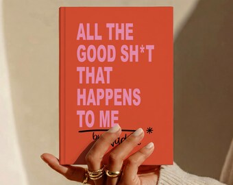 May include: A rectangular book with a vibrant orange cover featuring the text ALL THE GOOD SH*T THAT HAPPENS TO ME in bold pink capital letters. Below the text is a black horizontal line and a small black asterisk symbol on the right side.
