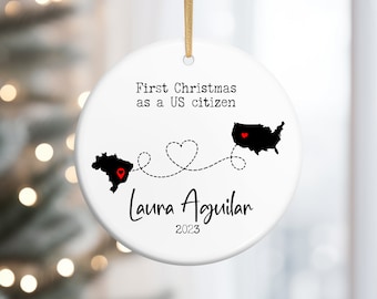 May include: White round Christmas ornament with black text that reads "First Christmas as a US citizen" and "Laura Aguilar 2023". The ornament features a black silhouette of the United States and Texas with red hearts in the center of each state.