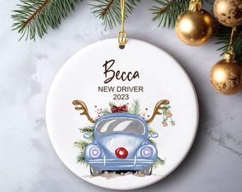 May include: A white ceramic ornament with a gold string for hanging. The ornament features a blue car with reindeer antlers and a red nose, surrounded by greenery. The text "Becca NEW DRIVER 2023" is printed on the ornament.