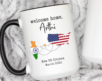 May include: White ceramic coffee mug with a black handle. The mug has a design of the United States and India with a heart connecting them. The text on the mug reads "welcome home, Arthi New US Citizen March 2024".
