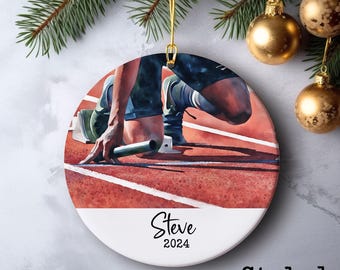 May include: Round ceramic ornament featuring a watercolor painting of a runner in a starting position on a red track. The ornament has the name "Steve" and the year "2024" printed on it. The words "Style 1" are printed at the bottom.