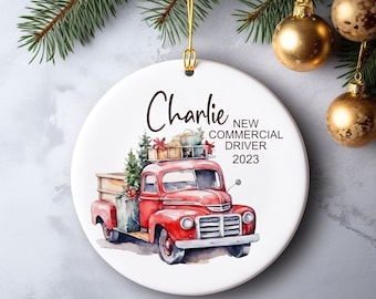 May include: A white ceramic Christmas ornament with a watercolor illustration of a red vintage truck carrying gifts and a Christmas tree. The ornament has the name "Charlie" and the text "NEW COMMERCIAL DRIVER 2023".
