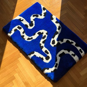 Op de afbeelding: Een blauw tapijt met een wit en zwart golvend patroon. Het tapijt is gemaakt van zacht, pluche materiaal en heeft een modern ontwerp.