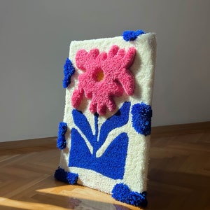 May include: A white rug with a pink flower and blue leaves. The rug is made of fluffy yarn and has a 3D effect.