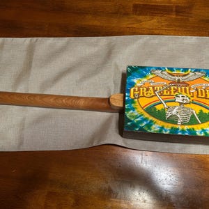 May include: A wooden cigar box guitar with a long, brown neck and a rectangular box featuring a colorful Grateful Dead design. The design includes a skeleton, eagle, and tie-dye background.
