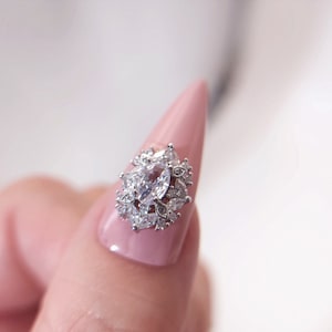 May include: A close-up of a fingernail with a silver and clear crystal nail art design. The design features a large, clear crystal in the center surrounded by smaller, clear crystals.